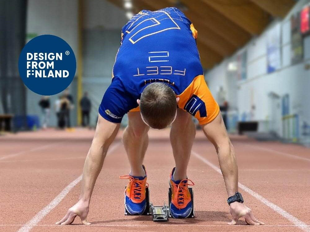 Person in a blue and orange outfit at the starting line of an indoor track, with 'Design from Finland' logo. 
Sini-oranssiin asuun pukeutunut henkilö sisäradan lähtöviivalla, ja hänen kuvassaan on 'Design from Finland' -logo.